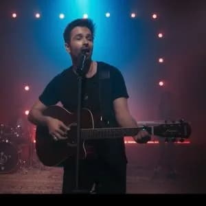 A singer playing an acoustic guitar into a microphone on a stage lit in red