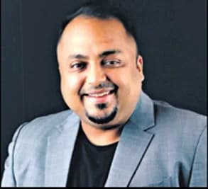 A portrait of a man in a grey blazer and black shirt, smiling at the camera