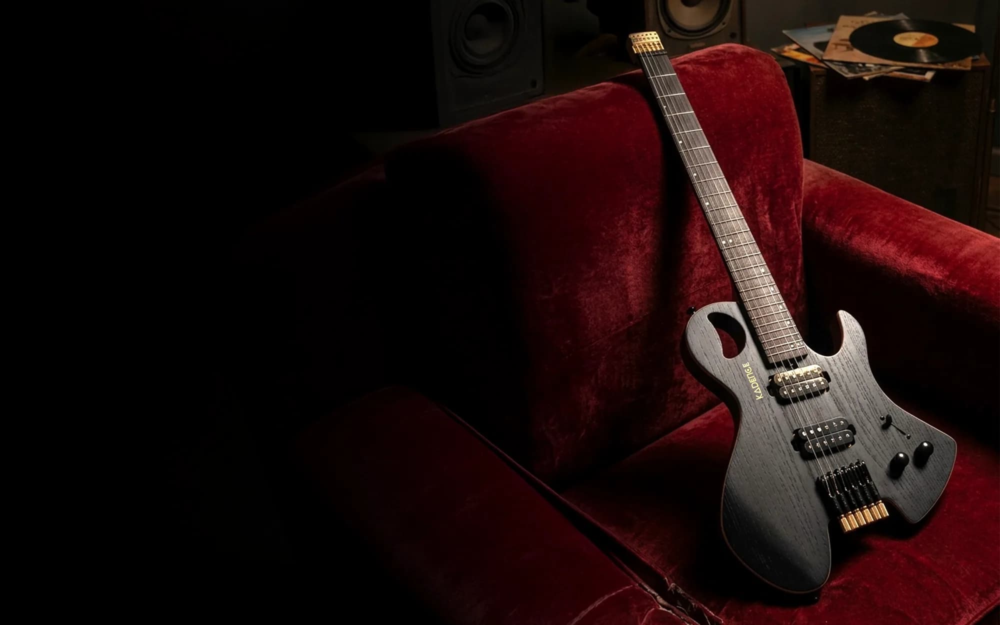 Black Kadence HL01 headless guitar leaning on a red velvet armchair, gold tuning knobs at the bridge and a gold clamp at the top of the neck, speakers and vinyl records behind it