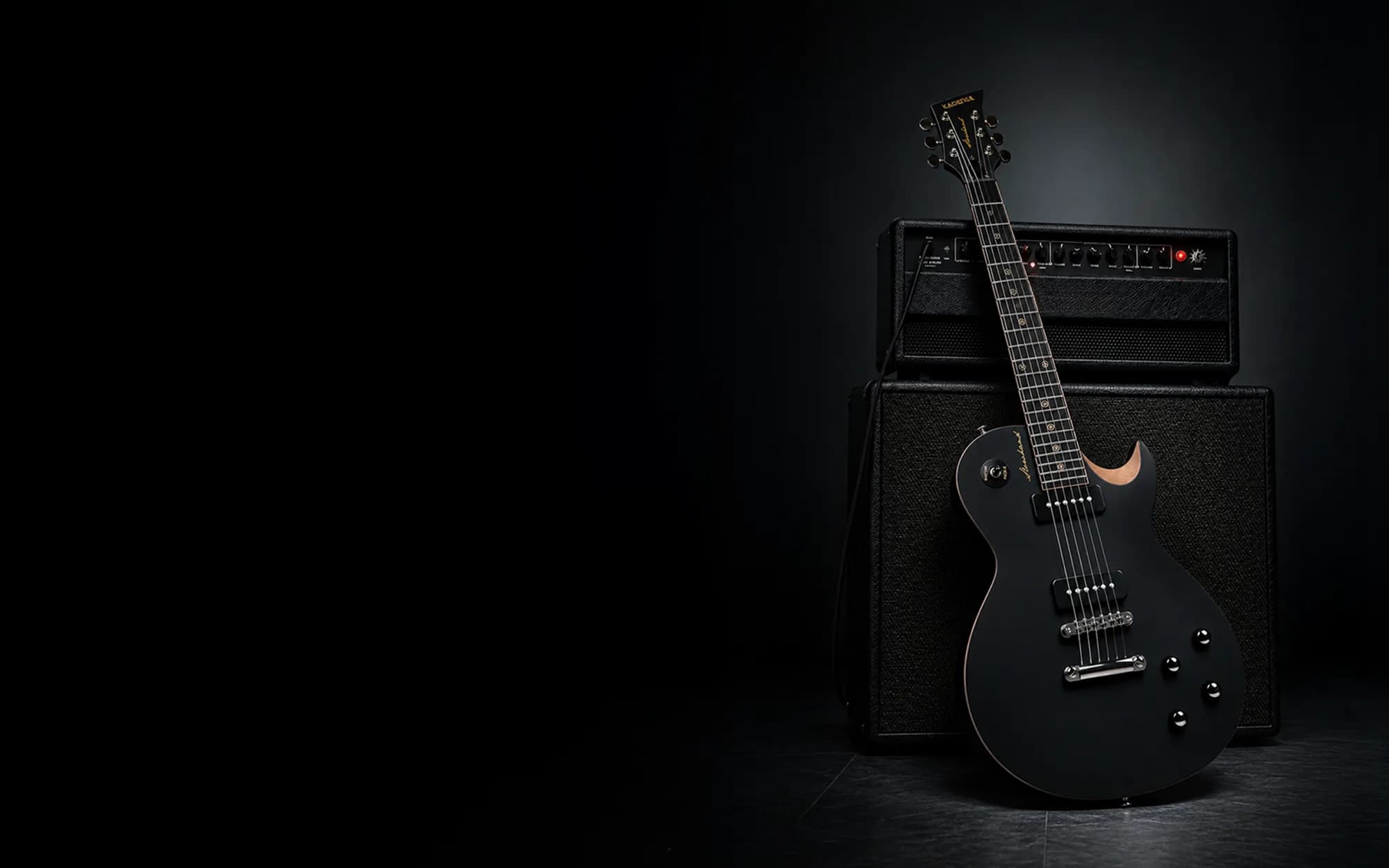 Kadence ESH04-P90 single-cutaway electric guitar in matte black with two P90 pickups, leaning against a black amp head and speaker cabinet in a dark room